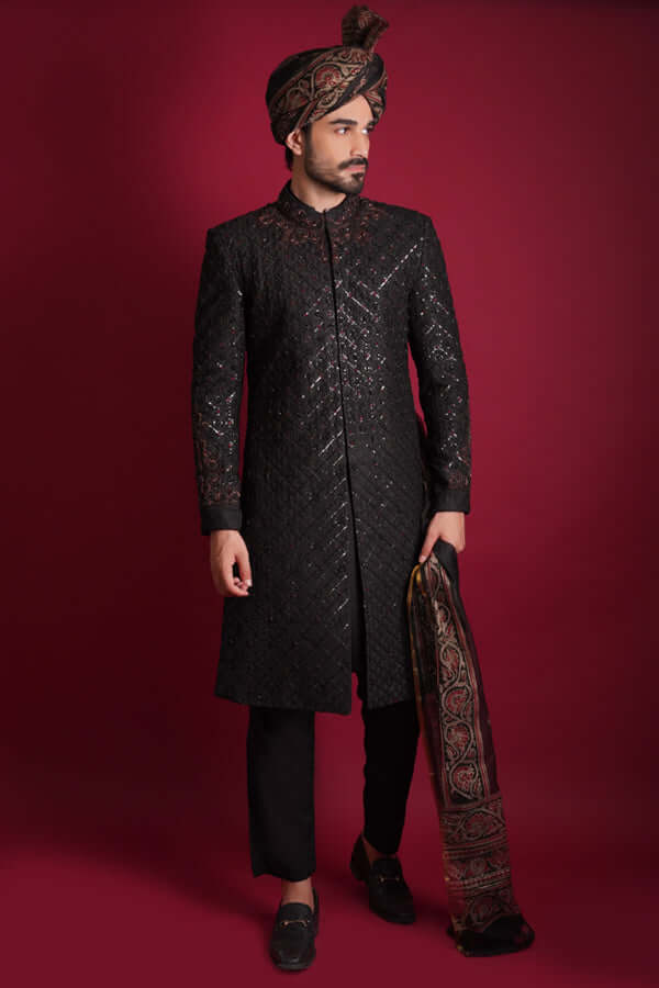 Full-length portrait of Arsalan Iqbal luxury black sherwani with a diamond-quilted pattern, featuring a mandarin collar with maroon embroidery, styled with a traditional Ajrak print shawl and a matching Ajrak turban.