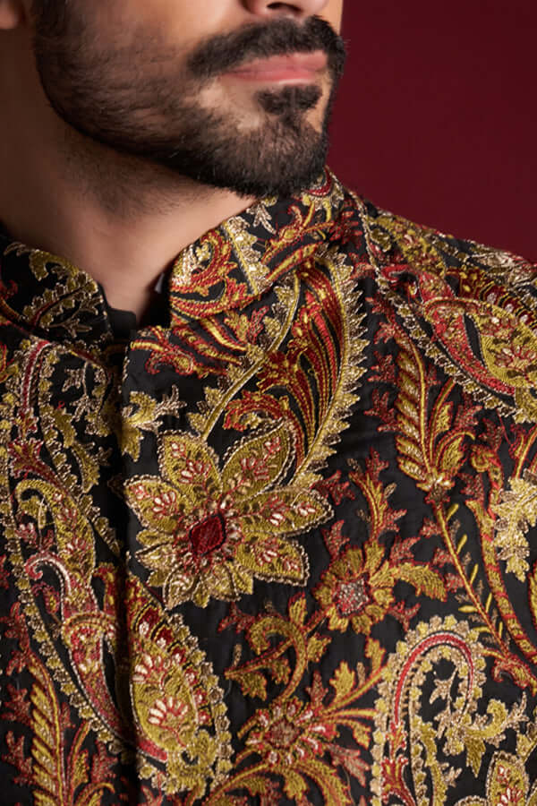 Extreme close-up of Arsalan Iqbal luxury silk sherwani featuring a rich amber, gold, and red paisley tapestry pattern with intricate gold thread embroidery and red bead accents.