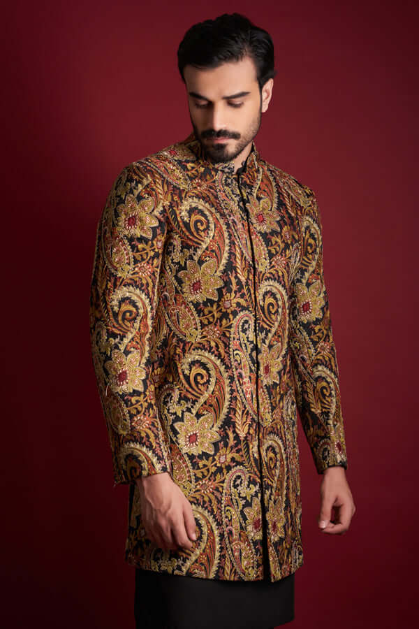A mid-length candid portrait of an Arsalan Iqbal luxury silk sherwani with a rich amber, gold, and red paisley tapestry print, featuring a mandarin collar and styled against a crimson background.