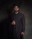 Front portrait of Arsalan Iqbal midnight black luxury sherwani featuring spaced-out tonal floral motifs, paired with a matching black turban and a heavily embroidered draped shawl.