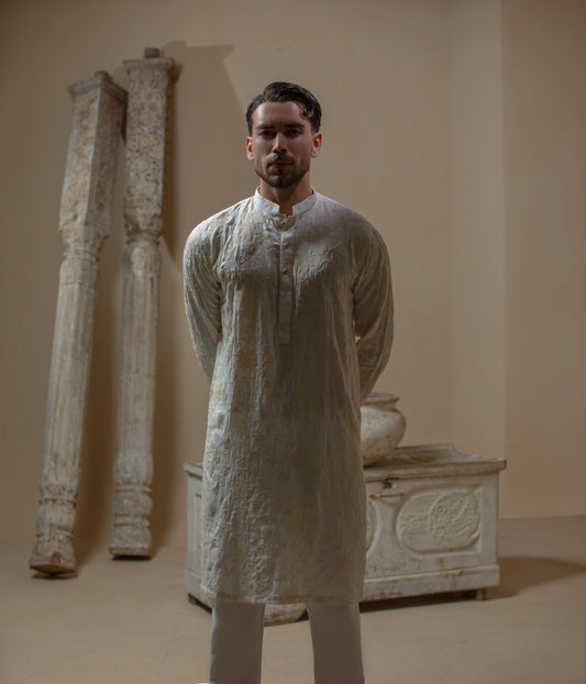 Man wearing Arsalan Iqbal's Cream Kurta Pyjama with allover gold and white floral embroidery, paired with white trousers and a watch, seated against a beige stone backdrop