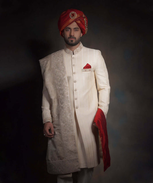 Arsalan Iqbal luxury cream sherwani featuring fine vertical linear textures, styled with a vibrant crimson red turban (kulla) with a jeweled brooch, a matching red pocket square, and an ivory embroidered shawl.
