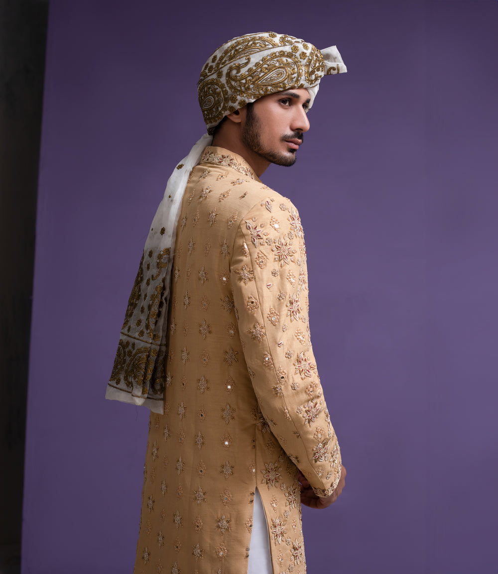 Side profile of an Arsalan Iqbal mustard gold designer sherwani featuring detailed floral embroidery and mirror-work, styled with a patterned turban and white trousers.