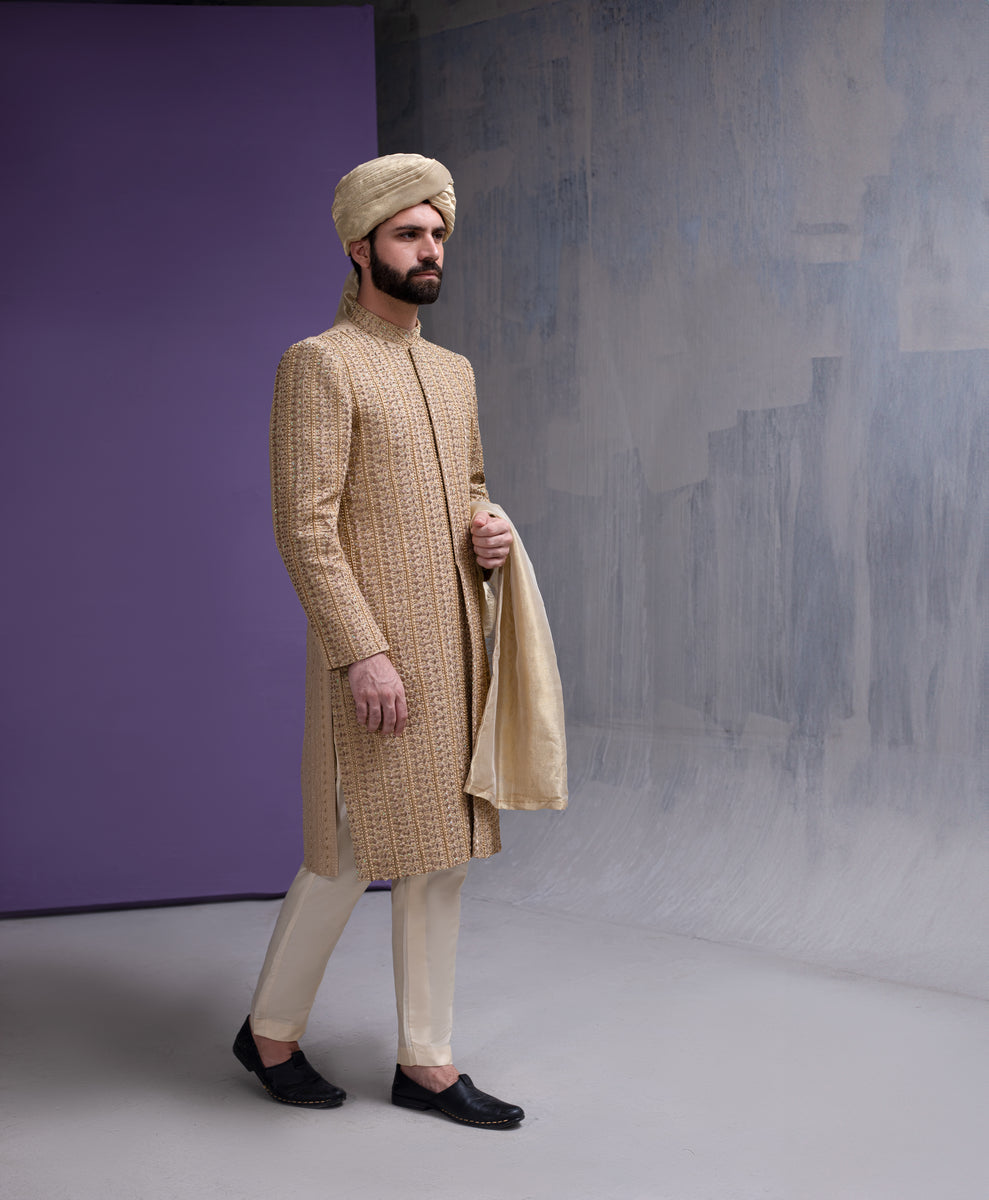 Full-length side profile of Arsalan Iqbal gold embroidered sherwani for men, paired with cream-colored trousers, black loafers, and a beige silk turban.