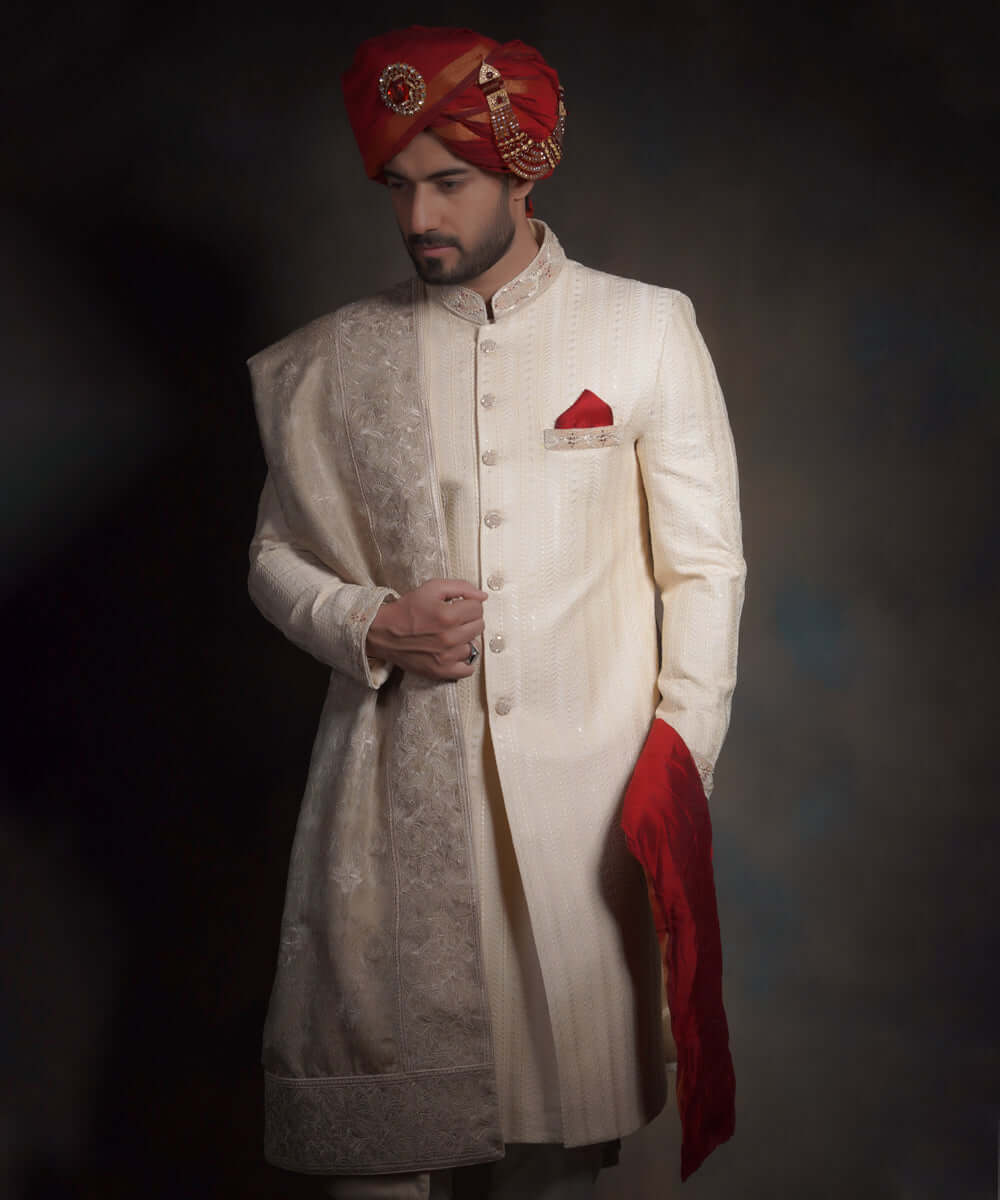 Side-profile portrait of Arsalan Iqbal heritage cream sherwani, featuring a vibrant crimson red kulla with a gold jeweled brooch, a red pocket square, and an intricately embroidered ivory shawl.