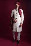 Full-length view of Arsalan Iqbal luxury ivory sherwani with self-on-self damask pattern, featuring embroidered collar and cuffs, styled with a maroon turban and matching silk shawl.
