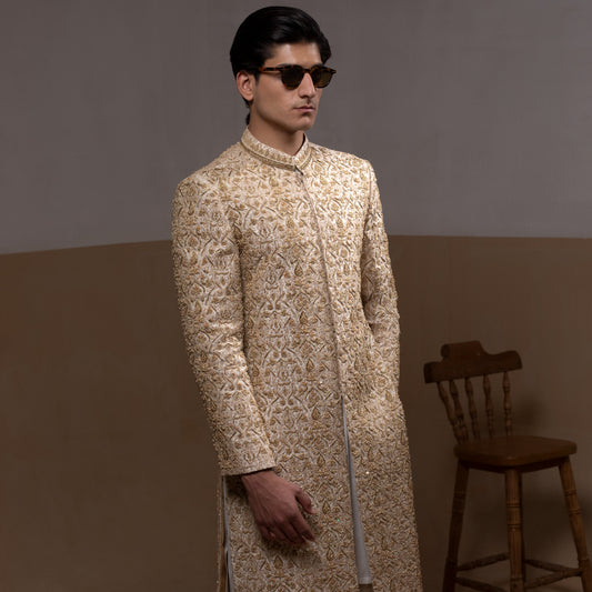 Arsalan Iqbal Ivory & Gold Handcrafted Maharaja Sherwani – man wearing a floor-length ivory sherwani with dense all-over gold hand embroidery featuring intricate floral and botanical motifs, mandarin collar, and a regal structured silhouette, accessorized with sunglasses, styled against a neutral two tone background with a wooden chair