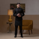 Arsalan Iqbal Jet Black Hand Embroidered Prince Jacket with Kurta Pyjama man wearing a fully embellished jet black prince jacket featuring dense hand embroidery with sequin and thread detailing, paired with a matching black kurta pyjama, mandarin collar, and a sleek formal silhouette, styled in a warm indoor setting