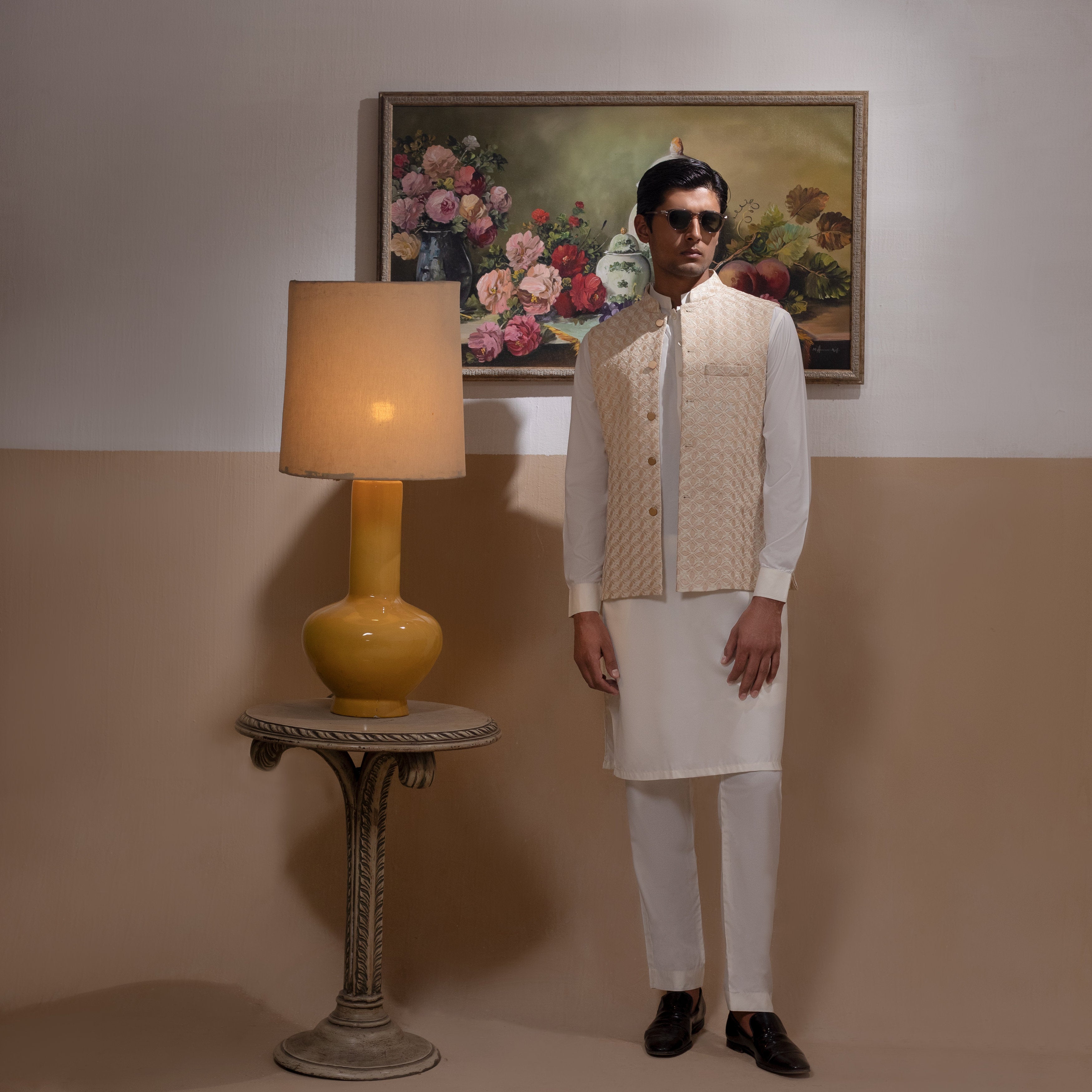 Arsalan Iqbal Light Gold Embroidered Waistcoat with IronEZ Kurta & Pyjama Set man wearing a light gold heavily embroidered sleeveless waistcoat with gold button closure over a crisp white full-sleeve kurta, paired with matching white straight-cut pyjama, accessorized with sunglasses, styled in a warm classic indoor setting