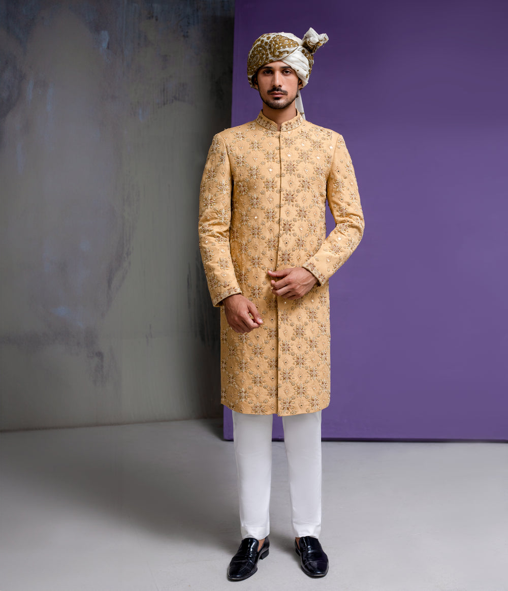 Arsalan Iqbal mustard gold groom sherwani with all-over floral embroidery and mirror-work highlights, paired with a white and gold patterned turban and white trousers.