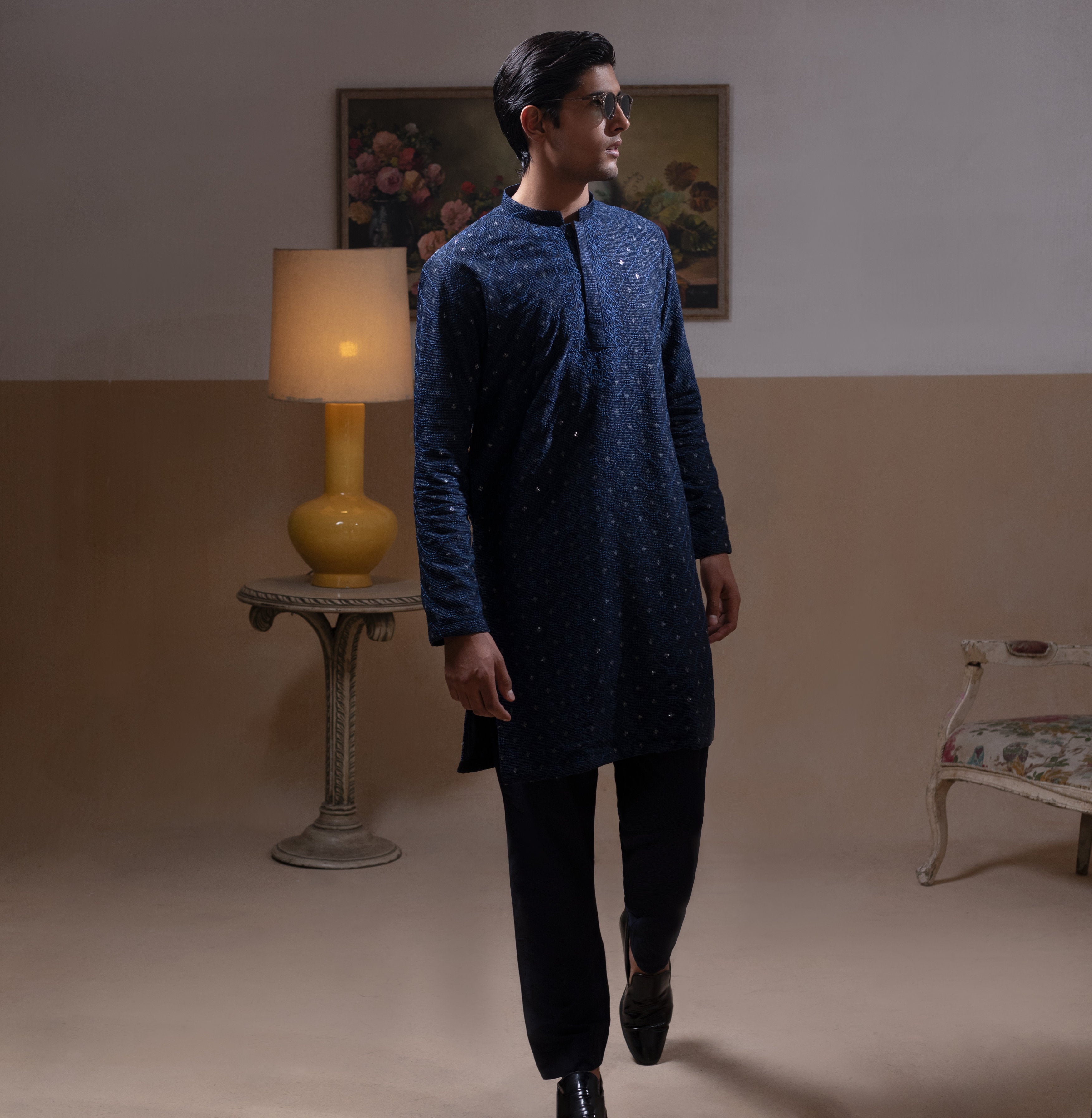 Man wearing Arsalan Iqbal's Navy Blue Allover Sequin Embroidered Kurta with dark navy trousers, black loafers and sunglasses, posed against a beige backdrop with yellow lamp and floral painting