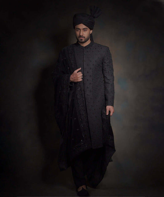 Arsalan Iqbal midnight black luxury sherwani for men, featuring spaced-out tonal floral motifs, a matching black turban with a high pleat, a draped velvet shawl, and black trousers.
