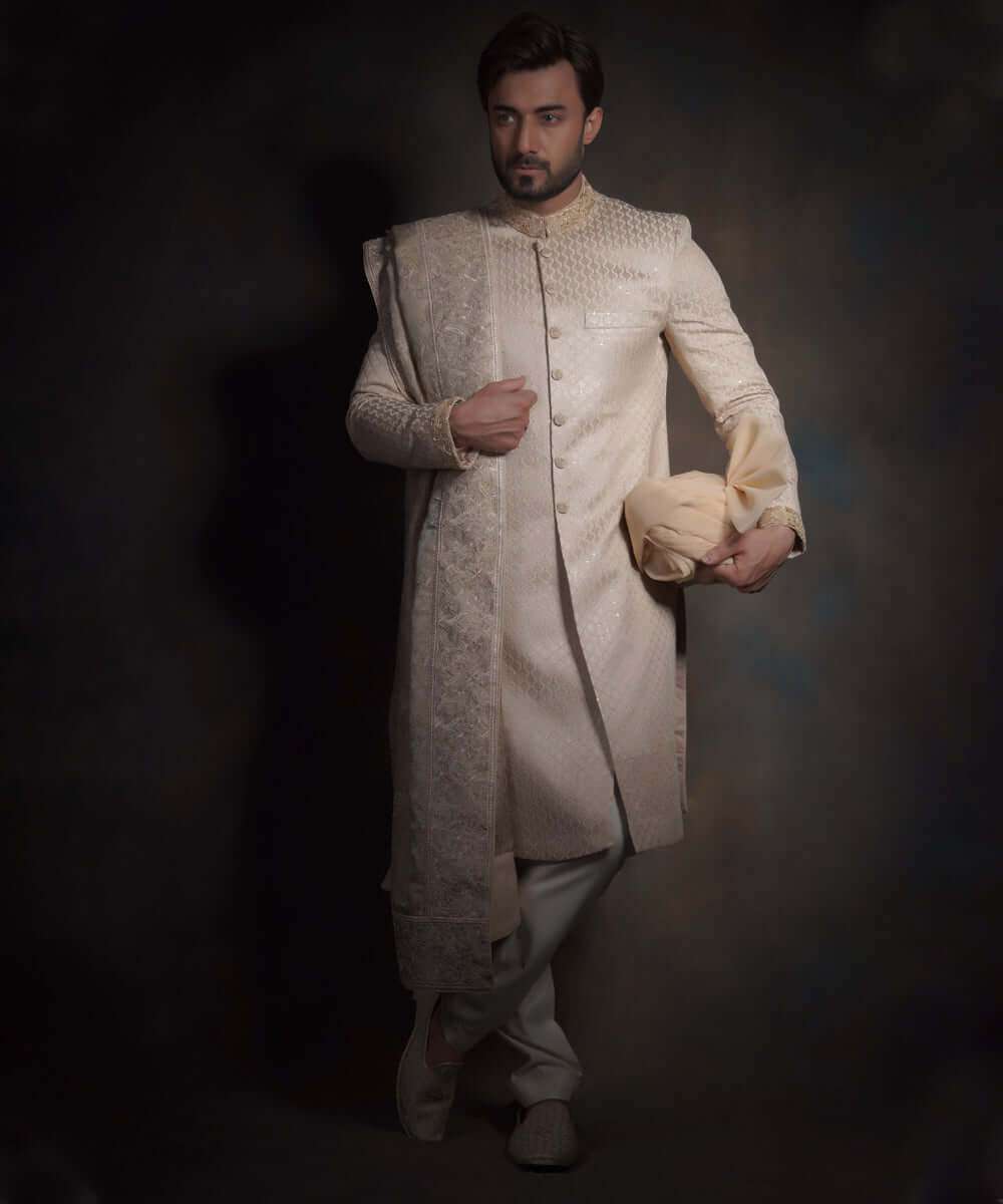 Full-length view of Arsalan Iqbal pearl cream quilted sherwani for grooms, featuring a diamond pattern, embroidered collar and cuffs, styled with a matching embroidered shawl, cream trousers, and traditional mojari shoes.