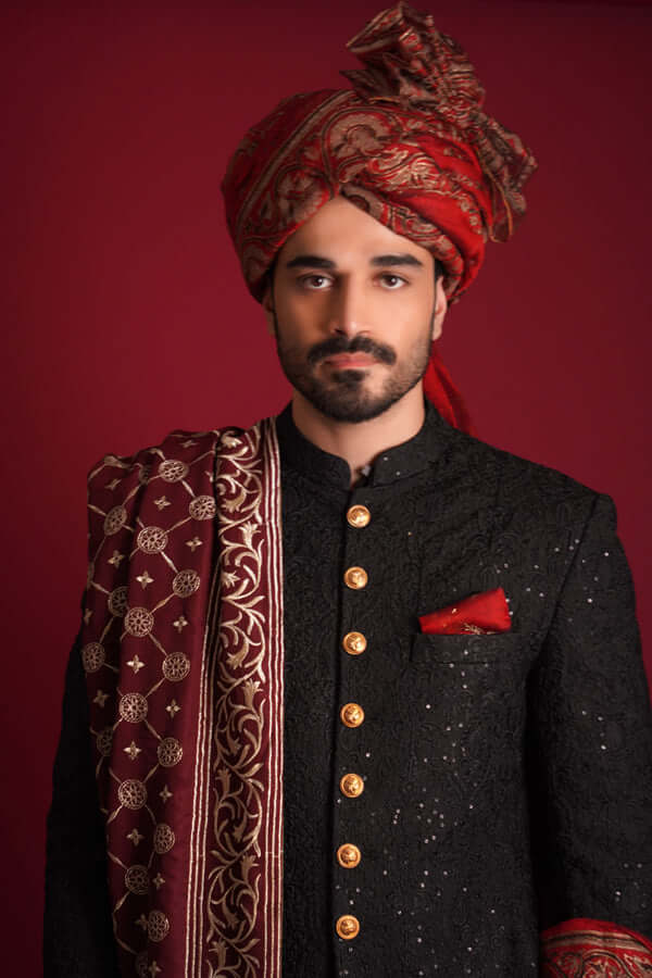 Mid-length portrait of Arsalan Iqbal luxury black sherwani featuring gold metallic buttons and a red silk pocket square, styled with a grand maroon silk turban and an intricately embroidered maroon shawl.