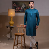Arsalan Iqbal Teal Blue Chikankari Kurta Pyjama man wearing a rich teal blue full-sleeve kurta with subtle all-over chikankari embroidery, mandarin collar with button placket, paired with white straight-cut pyjama, posed confidently with one hand on a wooden chair, styled in a warm classic indoor setting with a yellow vase and floral painting in the background
