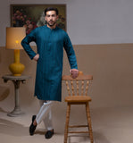 Arsalan Iqbal Teal Blue Chikankari Kurta Pyjama man wearing a rich teal blue full-sleeve kurta with subtle all-over chikankari embroidery, mandarin collar with button placket, paired with white straight-cut pyjama, posed confidently with one hand on a wooden chair, styled in a warm classic indoor setting with a yellow vase and floral painting in the background