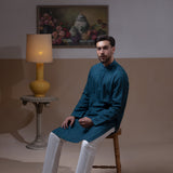 Arsalan Iqbal Teal Blue Chikankari Kurta Pyjama man wearing a rich teal blue full-sleeve kurta with subtle all-over chikankari embroidery, mandarin collar with button placket, paired with white straight-cut pyjama, posed confidently with one hand on a wooden chair, styled in a warm classic indoor setting with a yellow vase and floral painting in the background