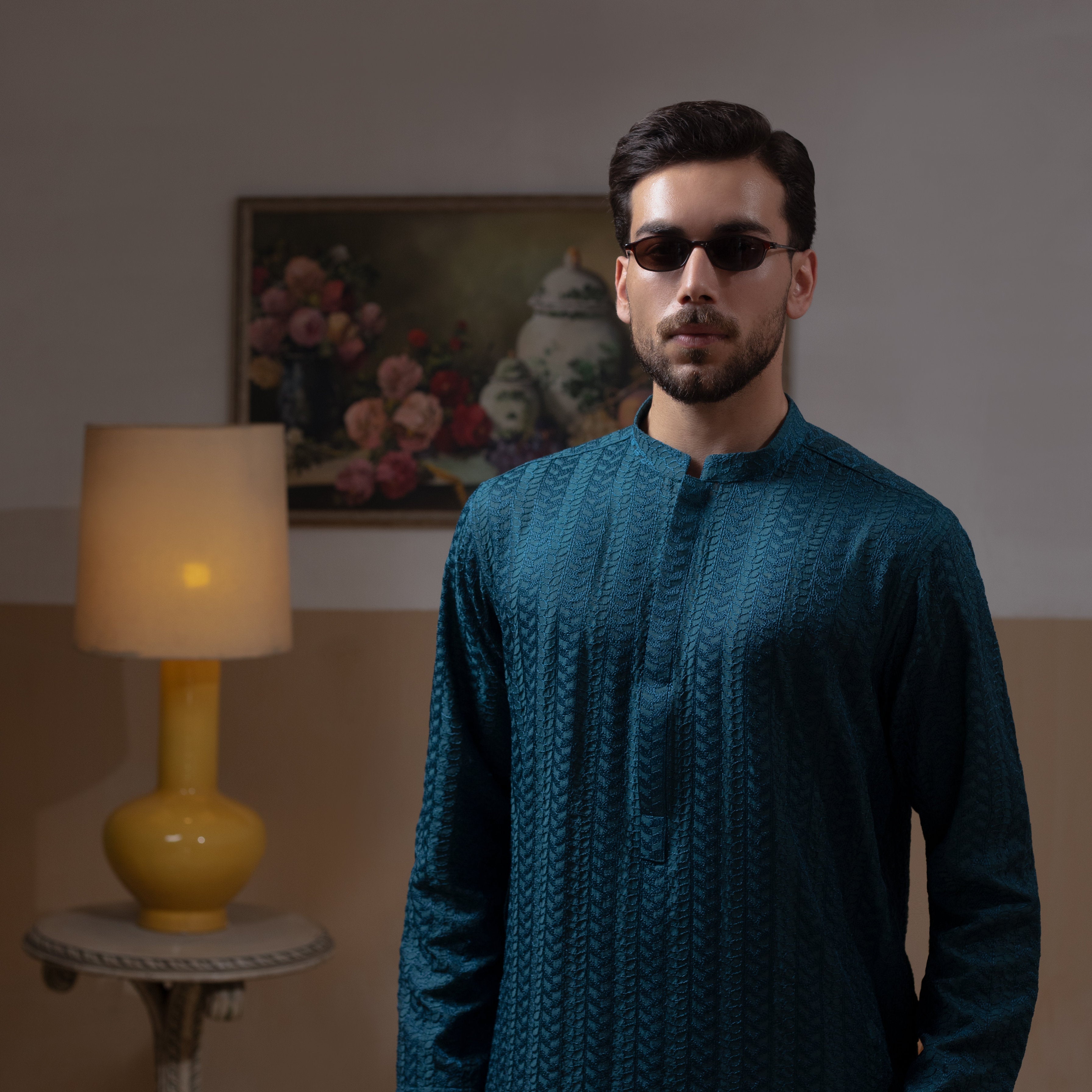 Arsalan Iqbal Teal Blue Chikankari Kurta Pyjama man wearing a rich teal blue full-sleeve kurta with subtle all-over chikankari embroidery, mandarin collar with button placket, paired with white straight-cut pyjama, posed confidently with one hand on a wooden chair, styled in a warm classic indoor setting with a yellow vase and floral painting in the background