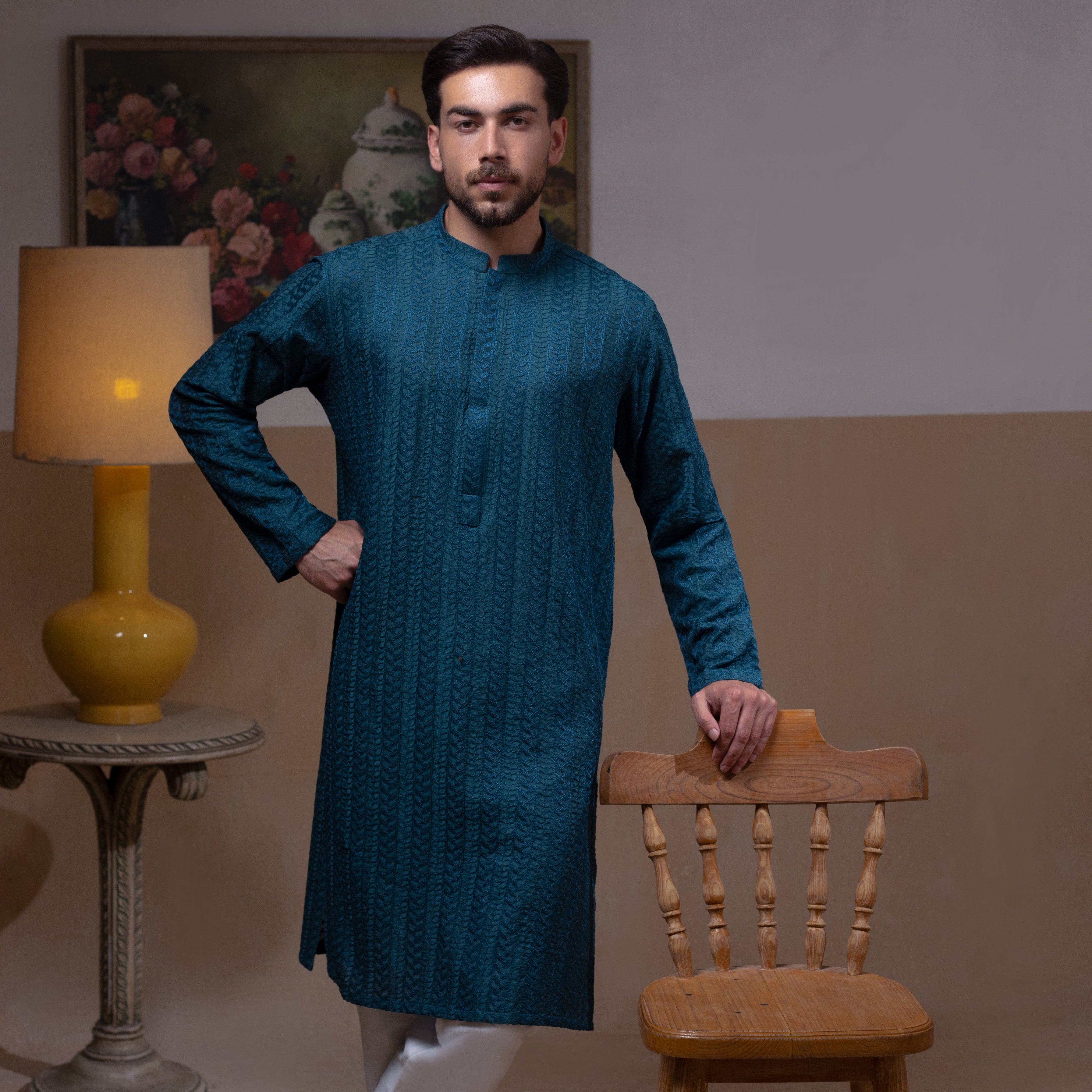 Arsalan Iqbal Teal Blue Chikankari Kurta Pyjama man wearing a rich teal blue full-sleeve kurta with subtle all-over chikankari embroidery, mandarin collar with button placket, paired with white straight-cut pyjama, posed confidently with one hand on a wooden chair, styled in a warm classic indoor setting with a yellow vase and floral painting in the background