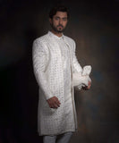 Portrait of Arsalan Iqbal snow white luxury sherwani for men, featuring geometric mirror-work, tonal embroidery, and a matching white turban with a row of decorative fabric buttons.