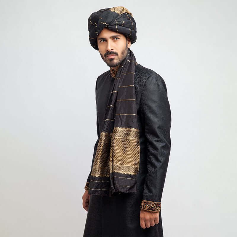 Medium portrait of a man in an Arsalan Iqbal black jacquard sherwani featuring embroidered cuffs and a black shawl with horizontal gold stripes and a patterned border.