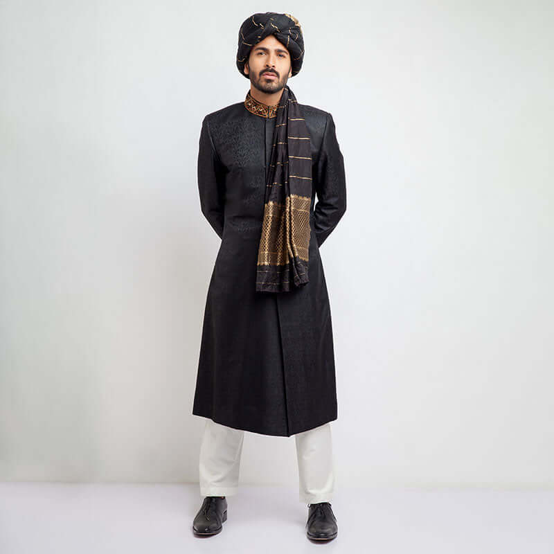 Full-length studio shot of a man in an Arsalan Iqbal black jacquard sherwani with an embroidered copper collar, paired with a black and gold striped turban and matching draped shawl.