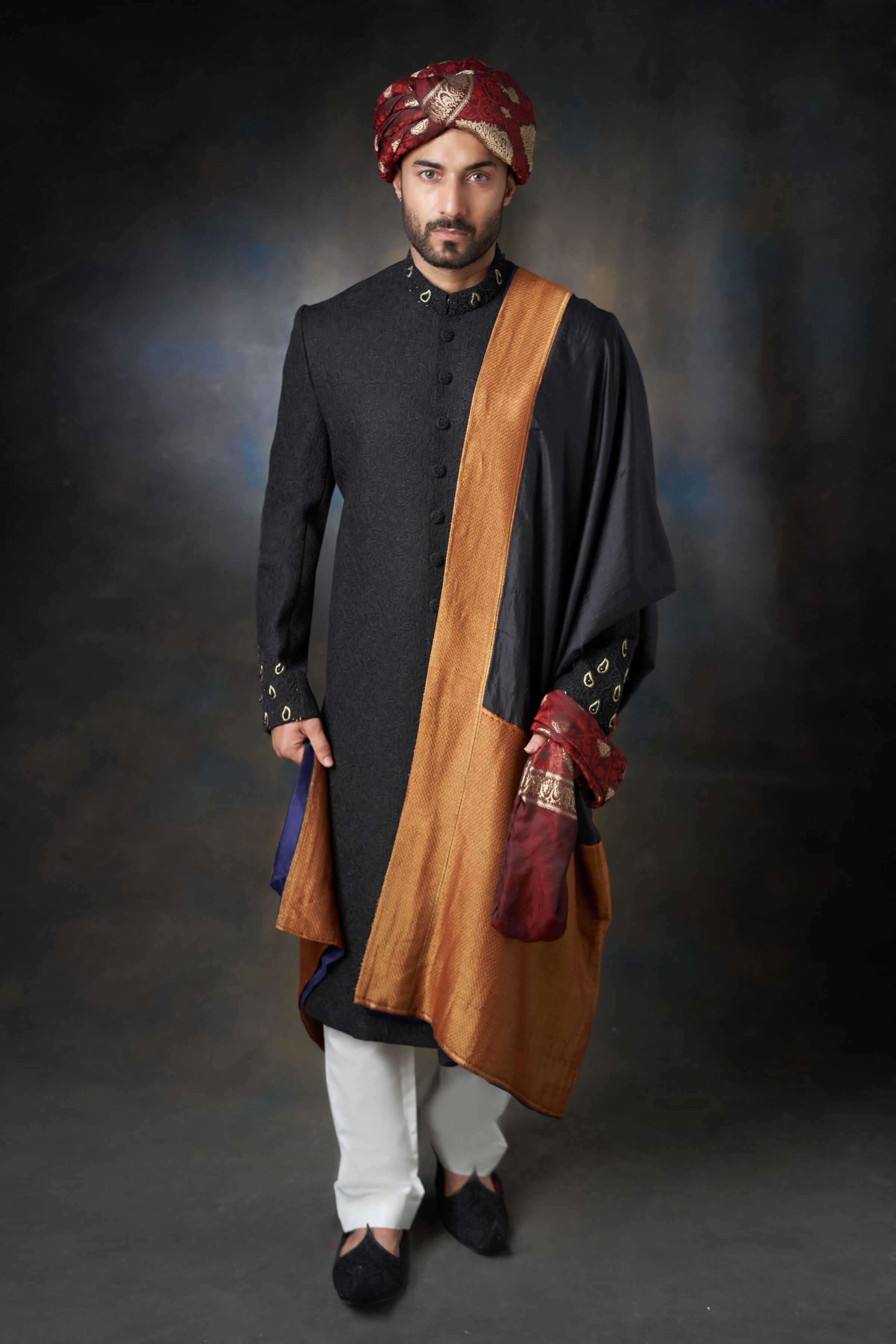 Man wearing an Arsalan Iqbal black sherwani with gold-bordered shawl, maroon patterned turban, and white trousers.