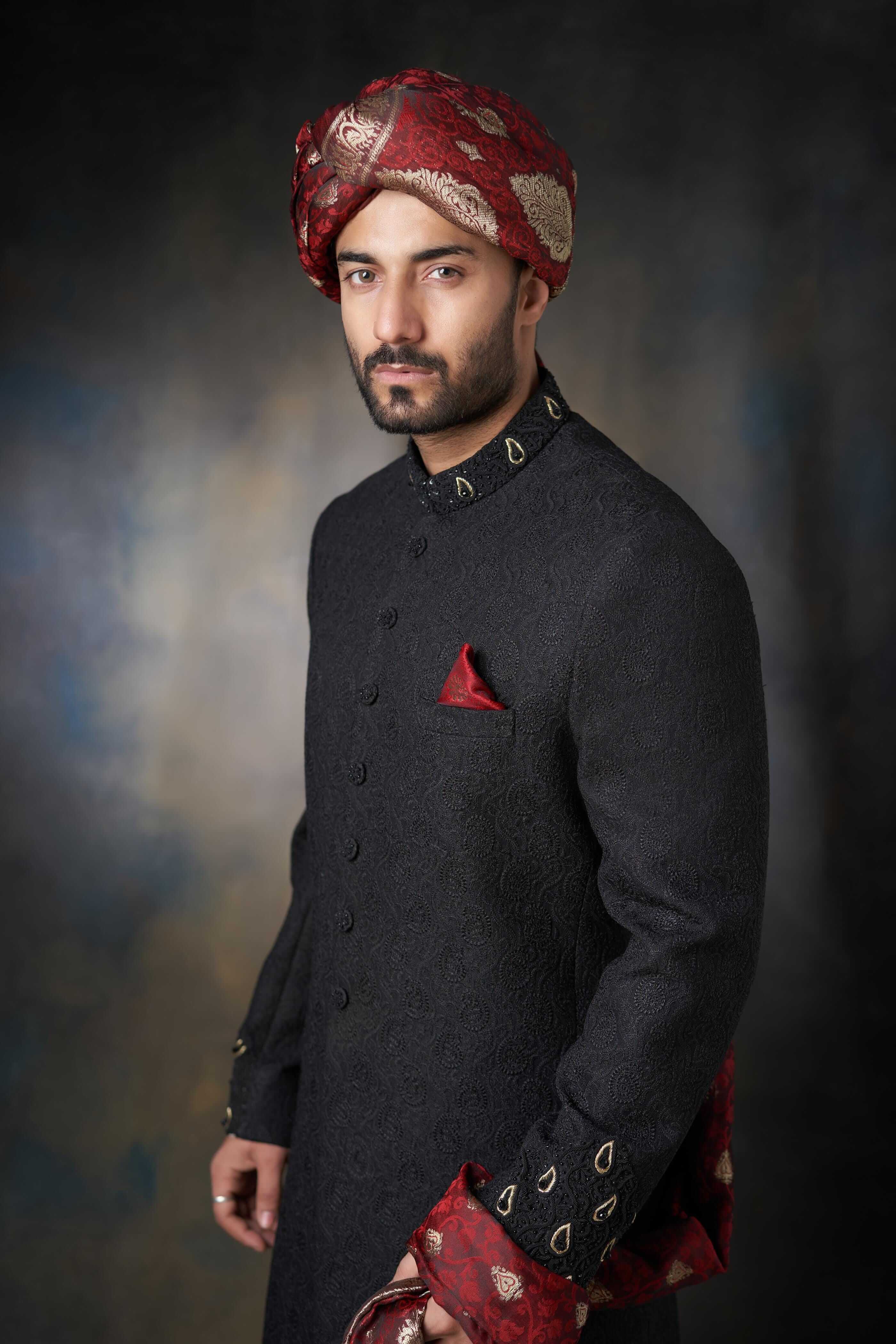 Full-length view of a man in an Arsalan Iqbal black textured sherwani with gold collar embroidery, maroon pocket square, matching turban, and white trousers.