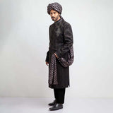 Three-quarter side profile of a man in an Arsalan Iqbal charcoal grey jacquard sherwani featuring silver chest embroidery and a draped silver polka dot silk shawl.