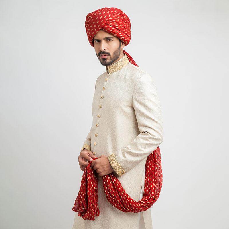 Side profile portrait of a man in an Arsalan Iqbal cream jamawar sherwani featuring an embroidered gold collar and a draped red silk shawl with gold polka dots.