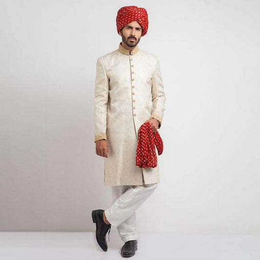 Full-length studio shot of a man in an Arsalan Iqbal cream jamawar sherwani with a gold embroidered collar and cuffs, paired with a vibrant red and gold polka dot turban and matching shawl.