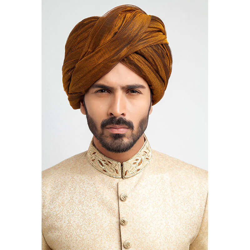 Full-length studio shot of a man in an Arsalan Iqbal gold jamawar sherwani with embroidered collar, mustard yellow pleated turban, and white trousers.