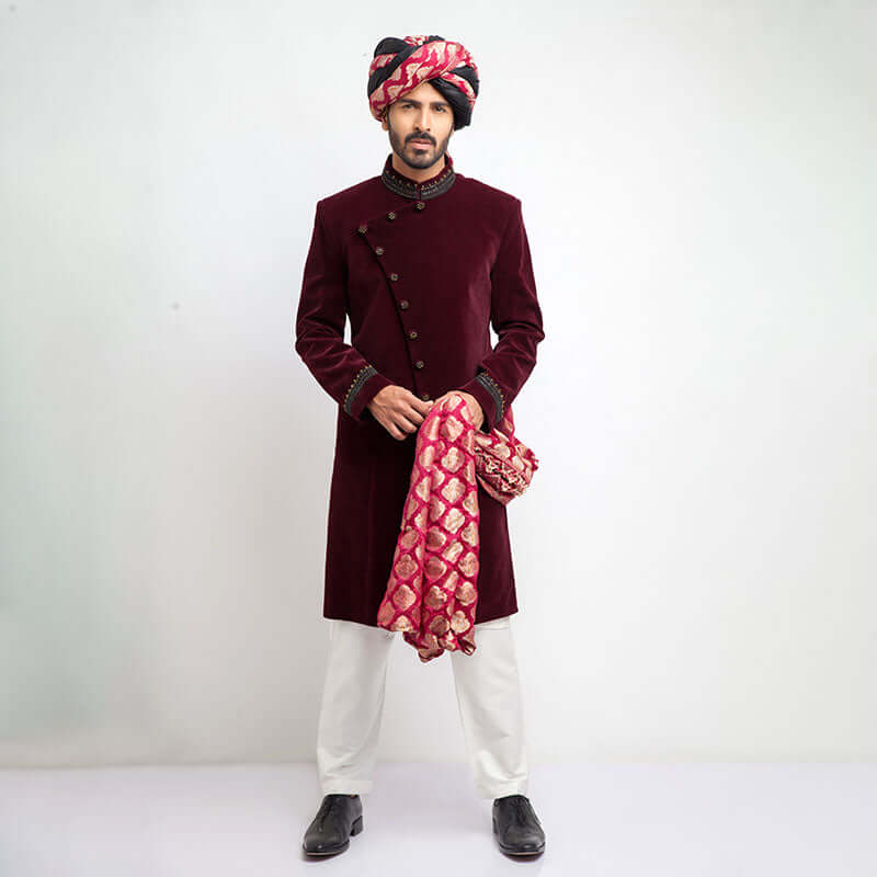 Full-length studio shot of a man in an Arsalan Iqbal maroon velvet sherwani with an asymmetrical button closure, paired with a pink and gold patterned brocade turban and matching shawl.