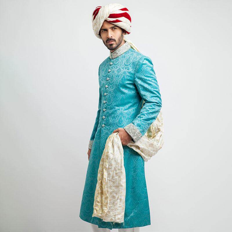 Full-length studio shot of a man in an Arsalan Iqbal teal jamawar sherwani with an all-over paisley pattern, featuring a silver embroidered collar and cuffs, paired with a white and maroon striped brocade turban.