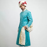 Full-length studio shot of a man in an Arsalan Iqbal teal jamawar sherwani with an all-over paisley pattern, featuring a silver embroidered collar and cuffs, paired with a white and maroon striped brocade turban.