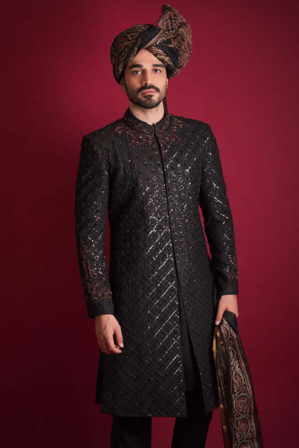 Arsalan Iqbal black quilted sherwani with subtle shimmer, featuring a maroon embroidered mandarin collar and cuffs, styled with a traditional Ajrak print turban and shawl.
