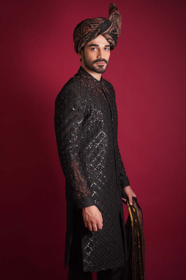 Side profile of Arsalan Iqbal luxury black quilted sherwani with red and silver beadwork, featuring an embroidered mandarin collar and styled with a traditional Ajrak turban against a red background.