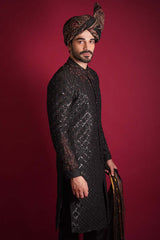 Side profile of Arsalan Iqbal luxury black quilted sherwani with red and silver beadwork, featuring an embroidered mandarin collar and styled with a traditional Ajrak turban against a red background.