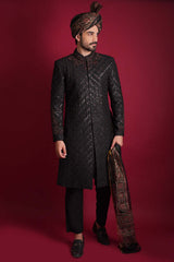 Full-length portrait of Arsalan Iqbal luxury black sherwani with a diamond-quilted pattern, featuring a mandarin collar with maroon embroidery, styled with a traditional Ajrak print shawl and a matching Ajrak turban.