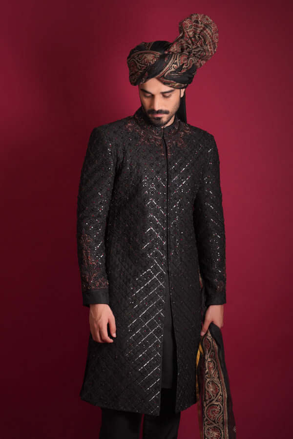A low-angle portrait of an Arsalan Iqbal luxury black quilted sherwani, featuring a maroon and black embroidered collar, styled with an Ajrak print turban and shawl against a crimson background.