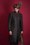 A low-angle portrait of an Arsalan Iqbal luxury black quilted sherwani, featuring a maroon and black embroidered collar, styled with an Ajrak print turban and shawl against a crimson background.