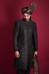 A low-angle portrait of an Arsalan Iqbal luxury black quilted sherwani, featuring a maroon and black embroidered collar, styled with an Ajrak print turban and shawl against a crimson background.