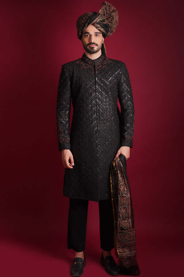 Full-length view of Arsalan Iqbal luxury black sherwani featuring a diamond-quilted pattern and subtle shimmer, styled with a traditional Ajrak print turban and matching Ajrak shawl, paired with black trousers and formal loafers.