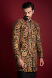 A mid-length candid portrait of an Arsalan Iqbal luxury silk sherwani with a rich amber, gold, and red paisley tapestry print, featuring a mandarin collar and styled against a crimson background.