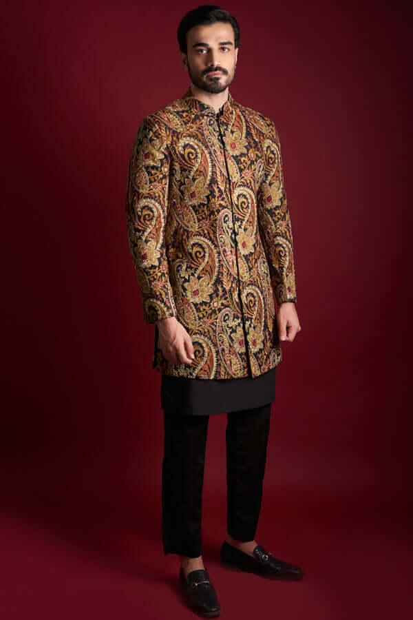 Full-length view of Arsalan Iqbal luxury short-length sherwani featuring a rich all-over amber, gold, and red paisley tapestry print on silk, styled with a black kurta-pyjama and formal loafers against a red background.