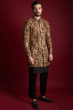 Full-length view of Arsalan Iqbal luxury short-length sherwani featuring a rich all-over amber, gold, and red paisley tapestry print on silk, styled with a black kurta-pyjama and formal loafers against a red background.