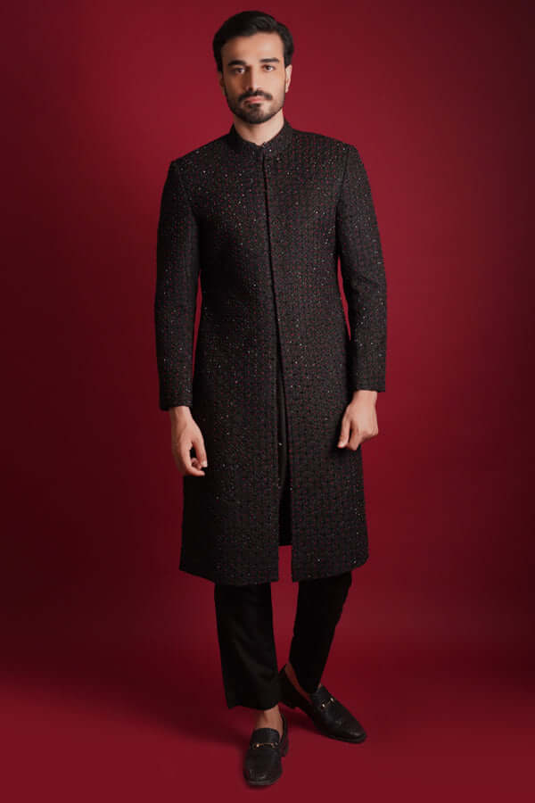 Full-length frontal view of Arsalan Iqbal luxury black sherwani featuring all-over maroon and green micro-embroidery, styled with black trousers and formal bit-loafers against a crimson background.