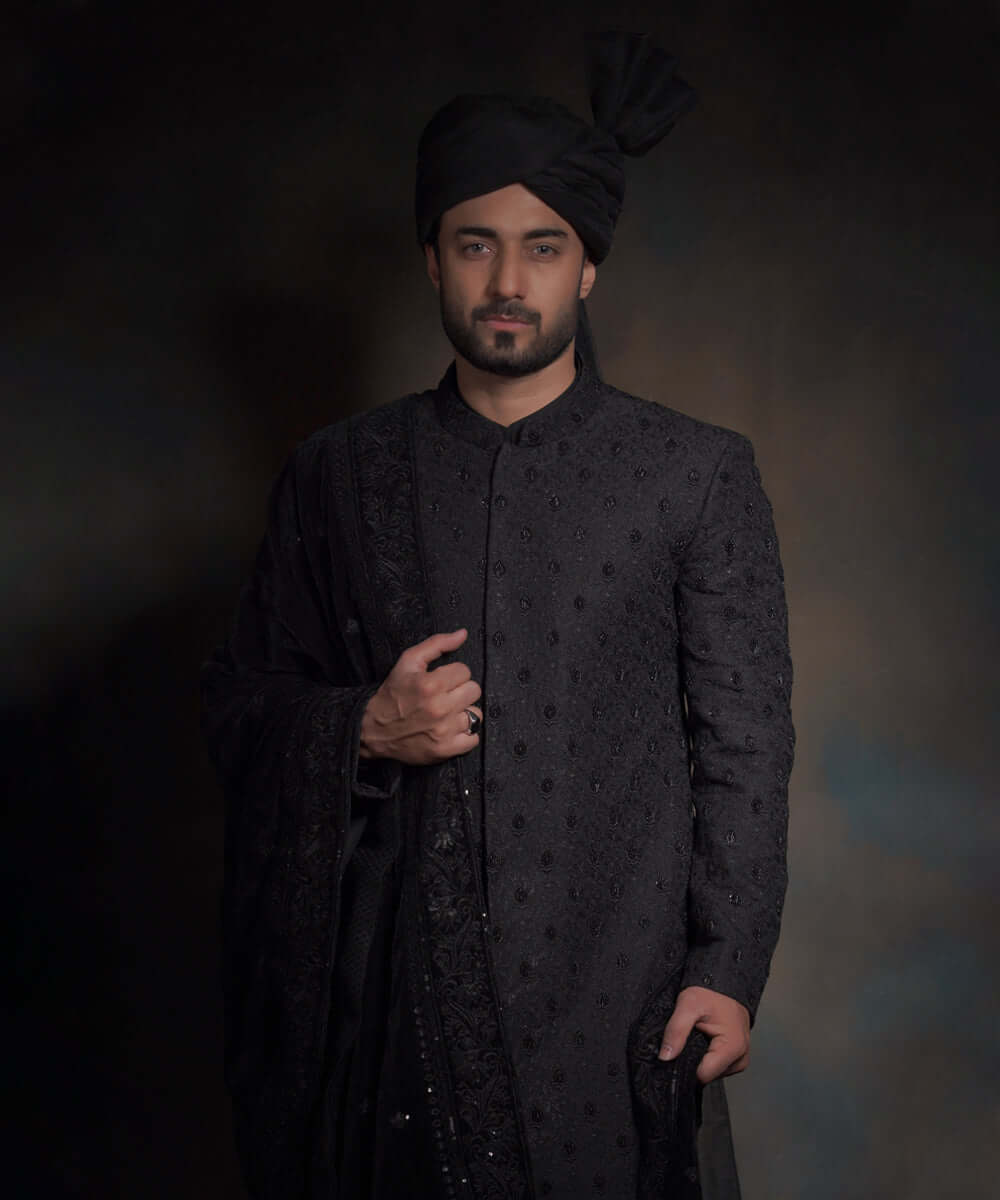 Front portrait of Arsalan Iqbal midnight black luxury sherwani featuring spaced-out tonal floral motifs, paired with a matching black turban and a heavily embroidered draped shawl.