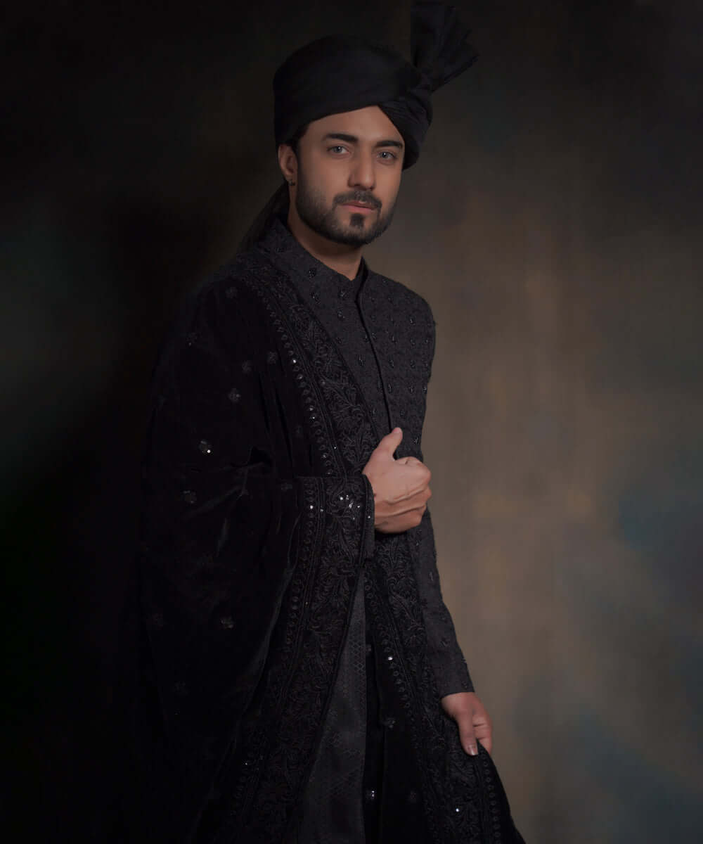 Portrait of Arsalan Iqbal midnight black sherwani with spaced-out tonal floral motifs, featuring a draped velvet shawl with heavy embroidery and a matching high-pleated turban.