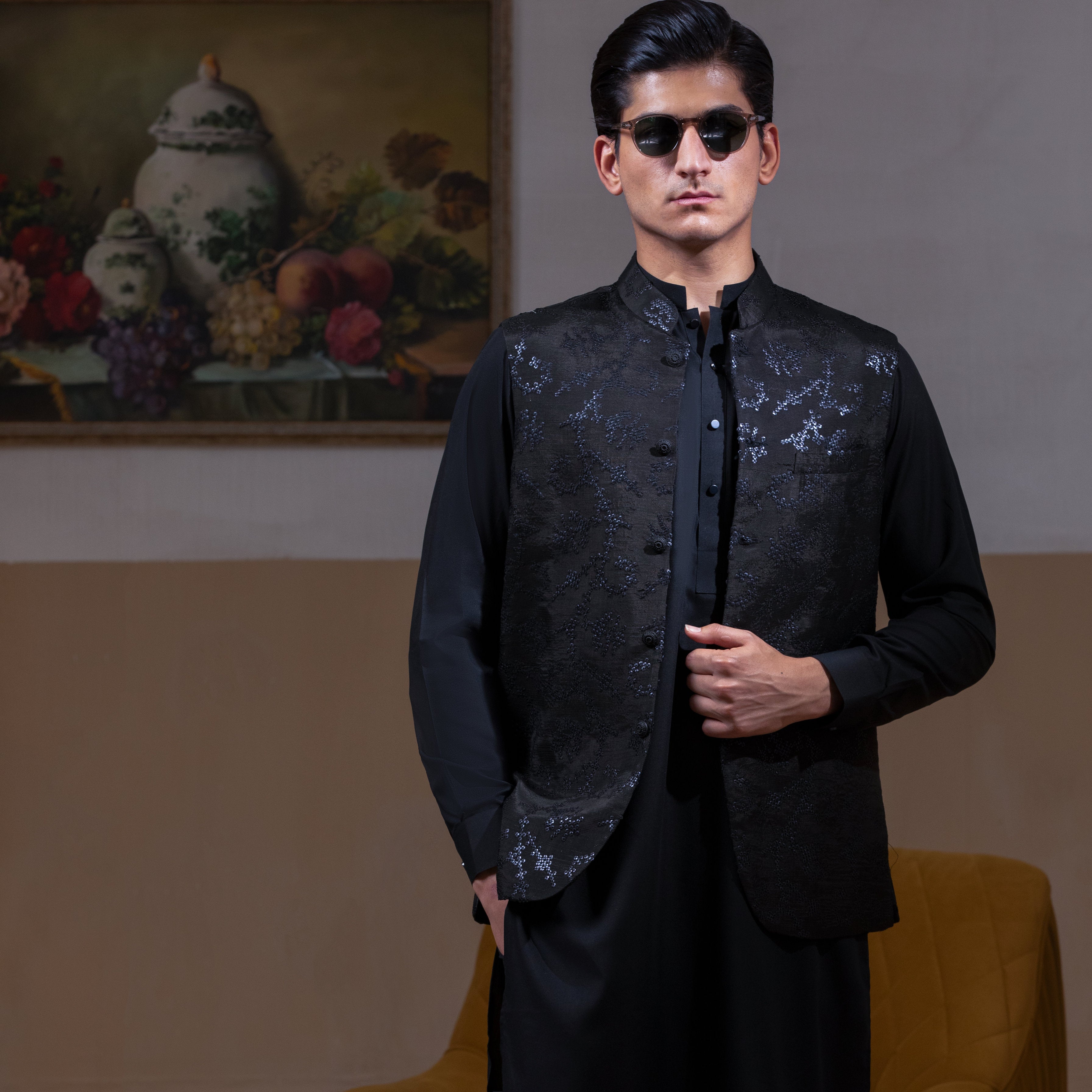 Man wearing Arsalan Iqbal's Black Sequin Embroidered Waistcoat over a matching black kurta pyjama, styled with embellished loafers and sunglasses, posed beside a mustard chair against a beige backdrop